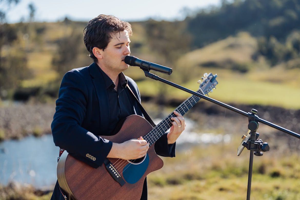 Harry Morris singing and playing acoustic guitar beside a creek at an outdoor wedding ceremony on a private farm