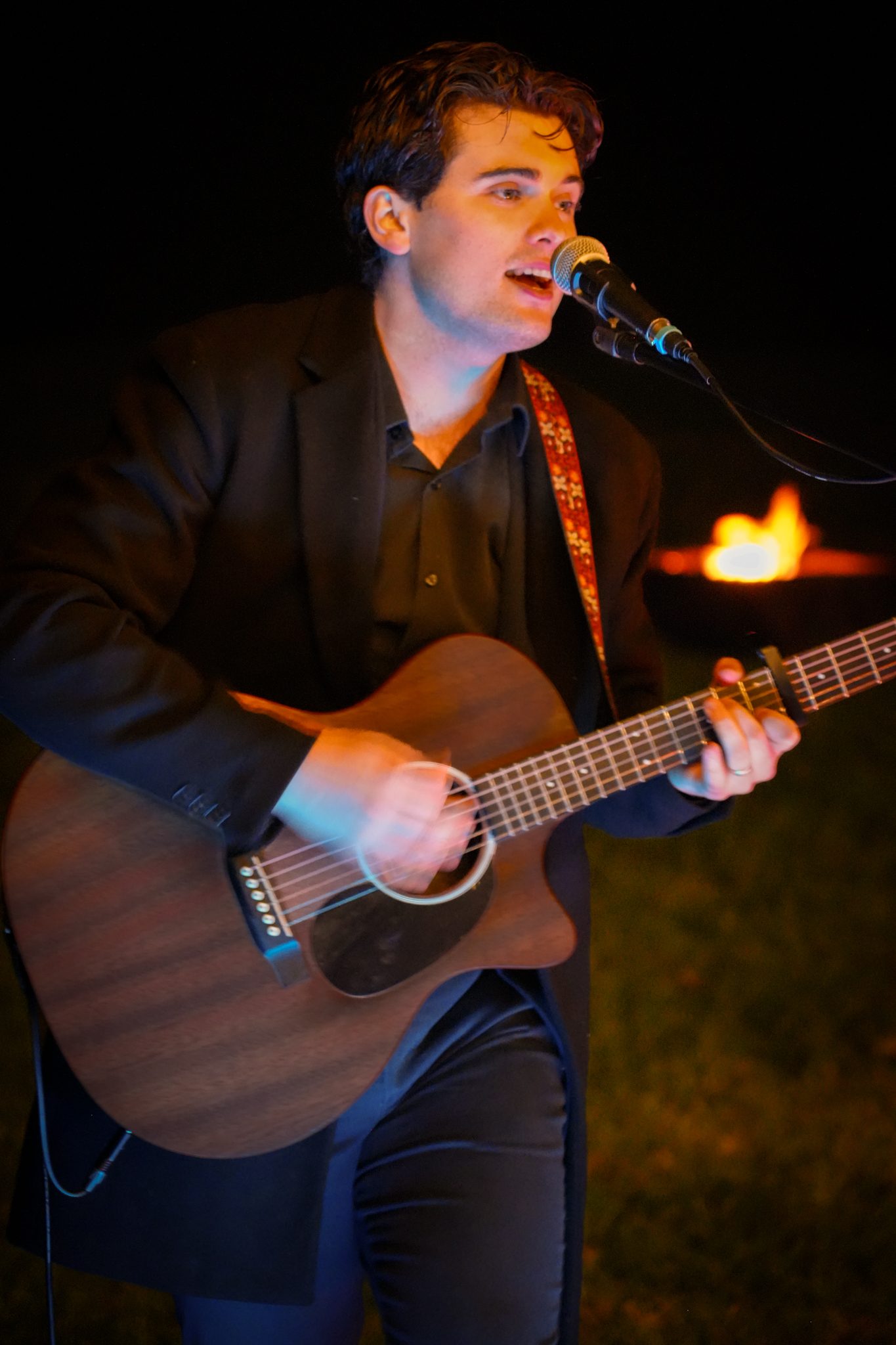Harry Morris singing and playing acoustic guitar at night, a fire pit burning behind him